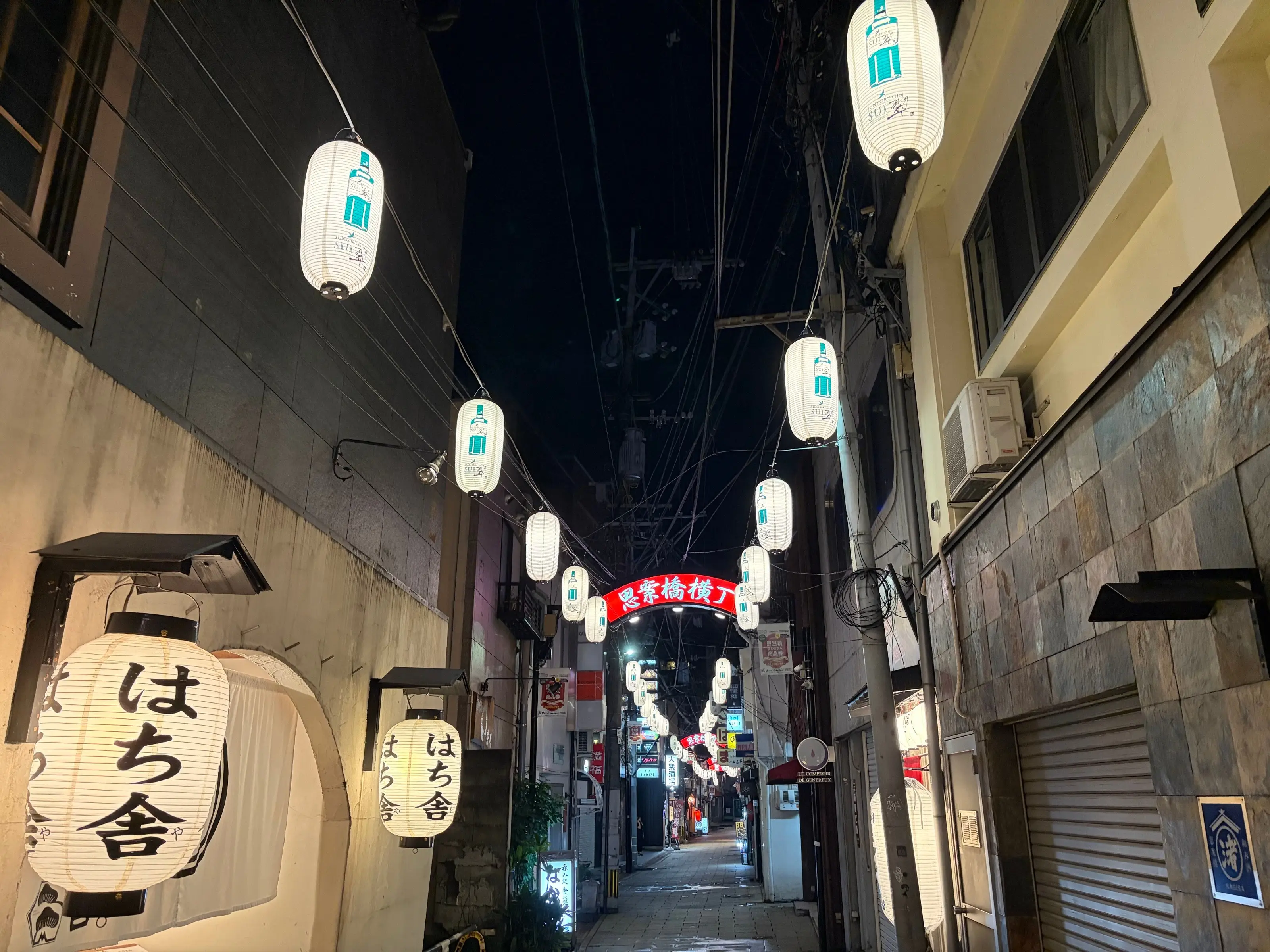 夜の石畳の路地。手前に「はち舎」の大きな提灯、奥まで白い提灯と赤い看板が続く